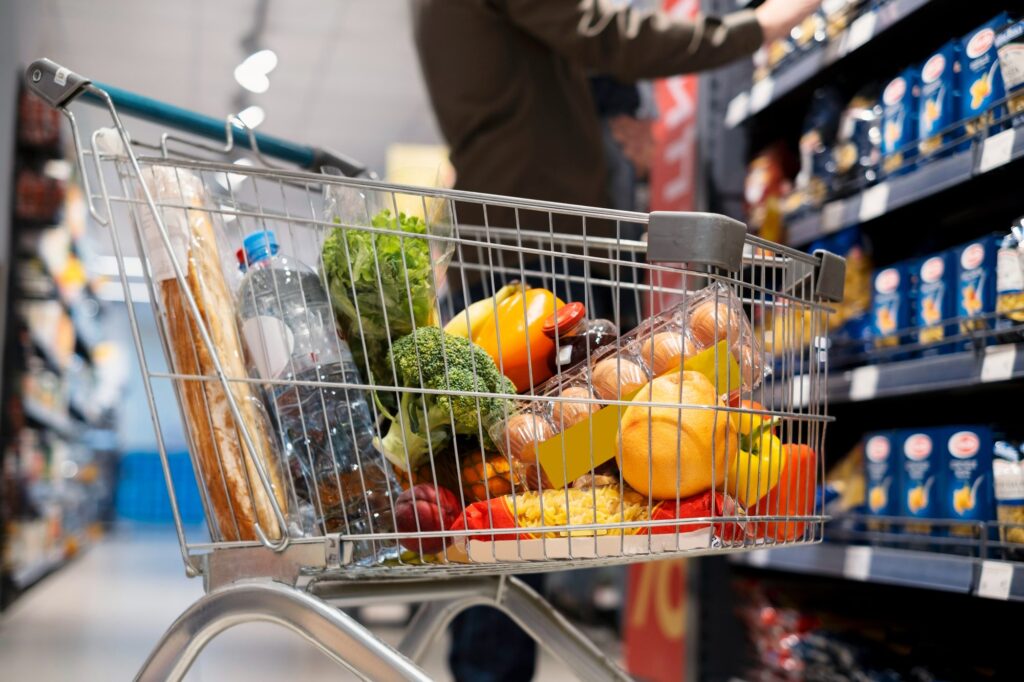 Carrinho de supermercado com frutas e verduras no corredor, ilustrando o consumo afetado pela Reforma Tributária