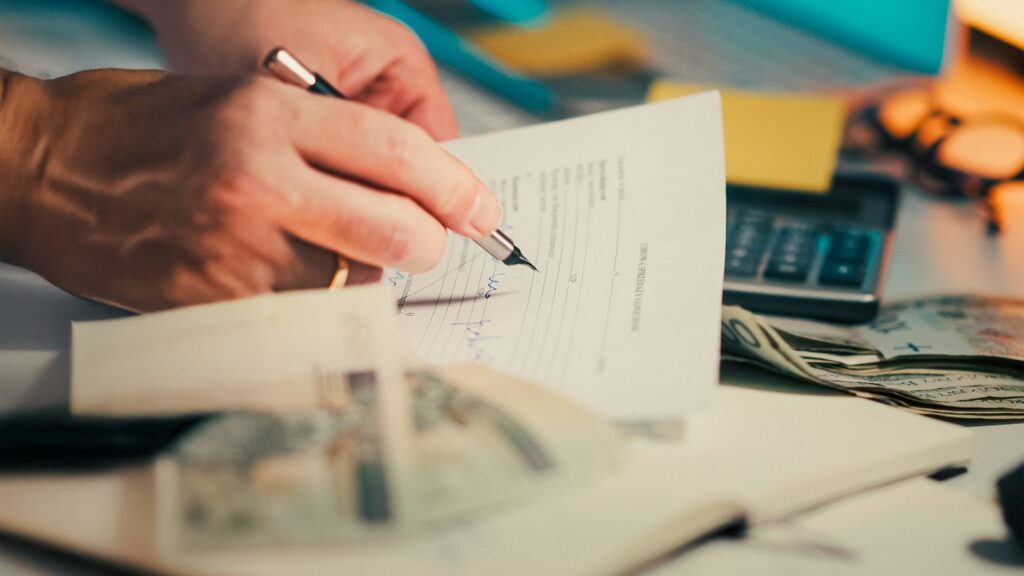 Mãos assinando documento sobre a mesa, representando a declaração do Imposto de Renda, a isenção até R$ 5 mil e o imposto mínimo